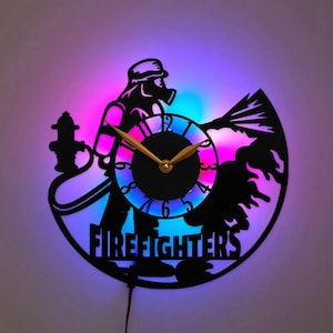 May include: A circular wall clock made from a black vinyl record featuring a silhouette of a firefighter with a hose and hydrant. The word FIREFIGHTERS is cut out at the bottom. The clock has gold hands and is illuminated by colorful LED lights.