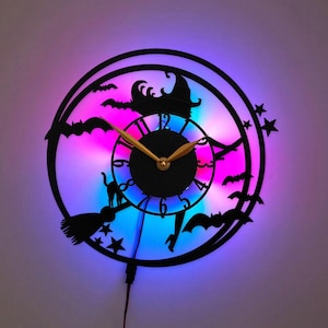 May include: A circular black wall clock featuring a silhouette of a witch on a broomstick, a cat, and bats against a vibrant purple and blue LED light background. The clock face has black numbers and gold-colored hands, with star cutouts.