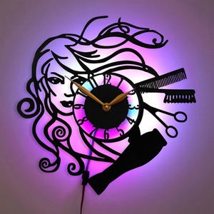 May include: A black metal wall clock with a silhouette of a woman with long hair, a hairdryer, scissors, and combs. The clock face has a black background with gold hands and a colorful ring around the edge. The clock is lit with multi-colored LED lights.