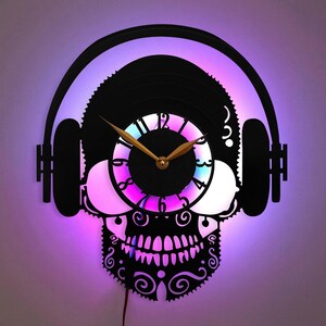 Könnte beinhalten: Schwarze, schädelartige Wanduhr mit Vinyl-Schallplatten-Design und Kopfhörern. Das Zifferblatt hat einen farbenfrohen, beleuchteten Farbverlauf. Die Zeiger der Uhr sind goldfarben. Die Uhr ist mit einem roten Kabel angeschlossen.