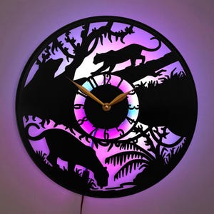 May include: Black vinyl record clock with a silhouette of two panthers in a jungle setting. The clock face is illuminated with a multi-colored LED light.