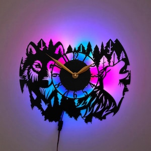 Può includere: Un orologio da parete nero con una silhouette tagliata al laser di un lupo e di un lupo che ulula su uno sfondo di foresta. Il quadrante ha lancette color oro ed è illuminato da una retroilluminazione a LED viola, blu e rosa.
