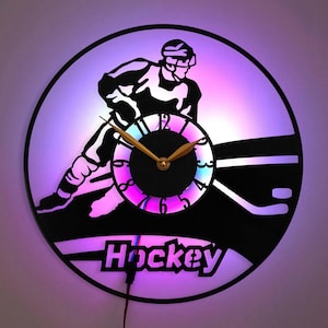 Puede incluir: Una silueta en blanco y negro de un jugador de hockey con un palo y un disco, con una esfera de reloj colorida y la palabra "Hockey" debajo. El reloj está iluminado con una luz morada y azul.