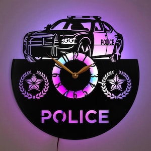 May include: Black vinyl record clock with a silhouette of a police car on top. The clock face has a rainbow-colored background and gold hands. The clock is surrounded by a black frame with the word "POLICE" in white letters and two star-shaped emblems.