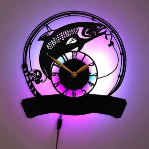 Puede incluir: Una silueta negra de un pez saltando fuera de un círculo de caña de pescar con una esfera de reloj en el centro. La esfera del reloj tiene manecillas doradas y un fondo de colores arcoíris. El reloj está iluminado con una luz morada y azul.