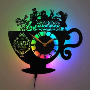 May include: A black wall clock shaped like a teacup with a silhouette of characters at a table. The text reads "We are all mad here" and "It's ALWAYS Tea time". The clock features gold hands and a vibrant, multi-colored LED backlight effect.