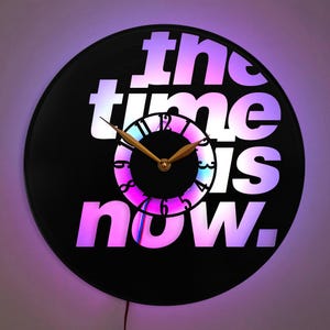Puede incluir: Un reloj de vinilo negro con letras blancas que dice "the time is now". El reloj tiene una aguja de hora y minuto de color dorado y una esfera de reloj colorida.