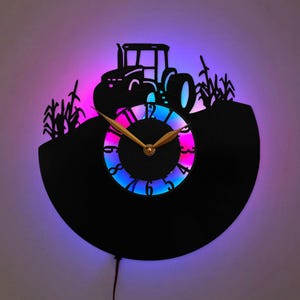 May include: A wall clock crafted from a black vinyl record featuring a silhouette of a tractor and corn stalks. The center dial has gold-colored hands and is illuminated by a vibrant LED light that shifts between shades of blue, purple, and pink.
