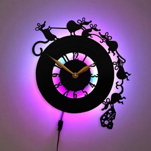 Pode incluir: Um relógio de vinil preto com ponteiro de hora e minuto dourado. O mostrador do relógio tem uma luz LED arco-íris atrás. O relógio tem a forma de um disco com uma silhueta de cinco ratos correndo ao redor da borda. Os ratos estão segurando um pedaço de queijo.
