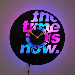 May include: A circular wall clock made from a black vinyl record featuring the text "the time is now." in a gradient of pink, purple, and blue light. The clock has a central black dial with black numbers and gold-colored hour and minute hands.