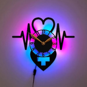 Könnte beinhalten: Eine schwarze Wanduhr mit einem zentralen runden Zifferblatt und goldenen Zeigern, umgeben von einer Herzschlaglinie, einem Herzsymbol oben und einem Herzsymbol unten mit einem Kreuz-Ausschnitt, beleuchtet durch blaues und rosa LED-Licht.