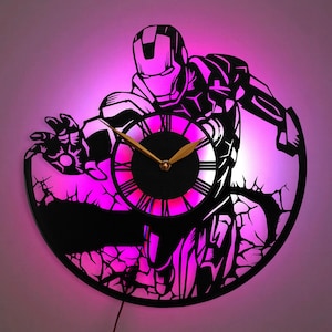Puede incluir: Un reloj de pared circular con la silueta negra de un superhéroe con armadura y la mano extendida. La esfera tiene números romanos y manecillas doradas. El diseño está retroiluminado con un resplandor LED de color rosa y violeta.