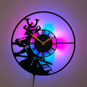 May include: A circular wall clock featuring a black silhouette of a samurai warrior. The clock face has gold-colored hands and black numerals. The design is illuminated from behind with a vibrant gradient of purple, pink, and blue LED light.