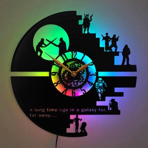 Op de afbeelding: Een ronde wandklok gemaakt van een zwarte vinylplaat met lasergesneden silhouetten van personages en een duel. De wijzerplaat toont cijfers en tandwielmotieven, verlicht door meerkleurige ledlampen met de tekst A long time ago in a galaxy far, far away....