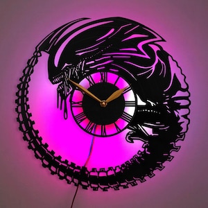 May include: A black and white wall clock with a silhouette of an alien creature. The clock has a pink LED light behind it.