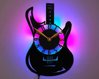 Könnte beinhalten: Eine schwarze Wanduhr in Form einer E-Gitarre mit einem runden Zifferblatt in der Mitte. Die Uhr hat goldfarbene Stunden- und Minutenzeiger und wird durch ein leuchtendes violettes und blaues LED-Licht hinter dem Rahmen beleuchtet.