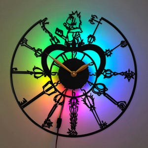May include: A circular wall clock featuring a black silhouette design of various key-shaped motifs and a central character figure. The clock face has gold-colored hands and is illuminated by a vibrant, multi-colored LED light background.