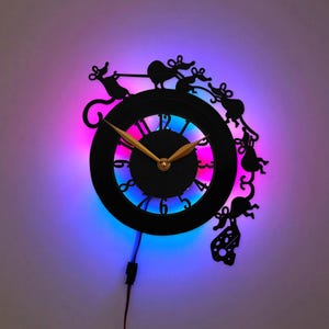 May include: A black wall clock featuring a circular face with gold-colored hands and cut-out numbers. The frame is adorned with silhouettes of multiple mice and a piece of Swiss cheese. The clock is illuminated by a vibrant purple and blue LED light.