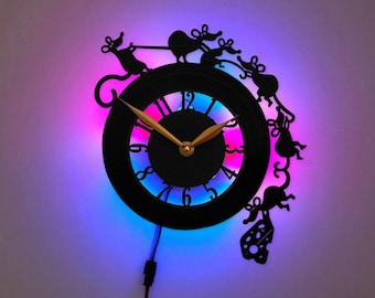 May include: A black wall clock featuring a circular face with gold-colored hands and cut-out numbers. The frame is adorned with silhouettes of multiple mice and a piece of Swiss cheese. The clock is illuminated by a vibrant purple and blue LED light.