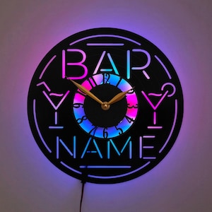 Può includere: Un orologio da parete circolare realizzato con un disco in vinile nero con un design ritagliato che riporta il testo BAR e NAME. Il quadrante è illuminato da una luce LED viola, blu e rosa, con due lancette color oro al centro.