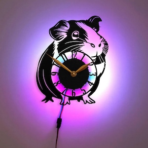 Könnte beinhalten: Eine schwarze Silhouette eines Meerschweinchens, das ein Zifferblatt mit bunten LED-Lichtern hält. Das Zifferblatt hat goldene Zeiger und schwarze römische Ziffern.
