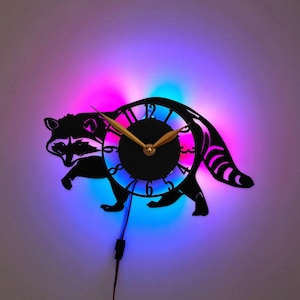 Pode incluir: Um relógio de parede preto em forma de silhueta de guaxinim com um mostrador circular central com ponteiros dourados e números pretos. O relógio é iluminado por trás por um vibrante display LED com tons de roxo, rosa e azul.