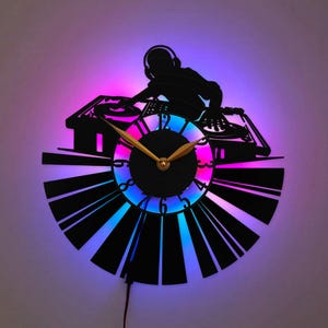 Peut inclure: Une horloge murale circulaire fabriquée à partir d'un disque vinyle noir représentant la silhouette d'un DJ à une table de mixage. Le cadran possède des aiguilles et des chiffres dorés, éclairés par une lueur LED violette et bleue.