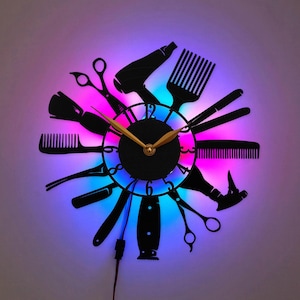 May include: A black wall clock featuring a circular face with gold hands and numbers. The frame is composed of silhouettes of hairdressing tools including scissors, combs, a hair dryer, and a razor. The clock is illuminated by a vibrant LED light.