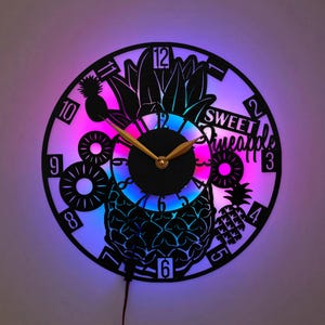 Op de afbeelding: Een ronde wandklok gemaakt van een zwarte vinylplaat met een uitgesneden ananasontwerp en de tekst SWEET Pineapple. De klok heeft goudkleurige wijzers en wordt van achteren verlicht met een levendige, meerkleurige LED-display.