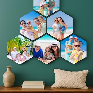 May include: A wall-mounted photo display featuring six hexagonal frames, each holding a beach vacation photo. The photos show families on a sunny beach. A wooden cabinet with a vase, books, and a pillow is in the foreground.