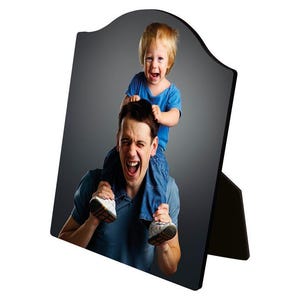 May include: A black wooden photo frame with a curved top and a stand. The frame displays a photograph of a man and a child. The man is wearing a blue shirt and the child is sitting on his shoulders, both with their mouths open.