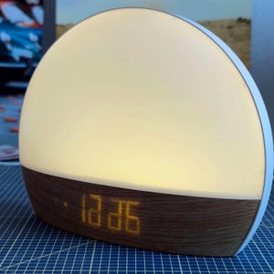 Pode incluir: Um despertador de nascer do sol branco e castanho com um visor digital mostrando a hora. O relógio tem uma forma arredondada com uma base de grão de madeira e um mostrador brilhante e iluminado.