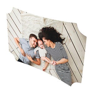 May include: A family of three lies on a light-colored wooden floor, with a white knitted blanket. The parents are wearing casual clothing, and the baby is in a white onesie. The mother wears a black and white striped dress.