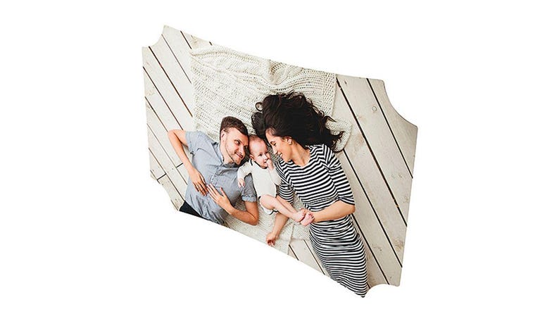 May include: A family of three lies on a light-colored wooden floor, with a white knitted blanket. The parents are wearing casual clothing, and the baby is in a white onesie. The mother wears a black and white striped dress.