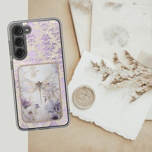 May include: A clear phone case featuring a lavender floral and script pattern. The case has a rectangular inset with a dragonfly and floral design in shades of lavender and white. The phone case is on a light brown surface.