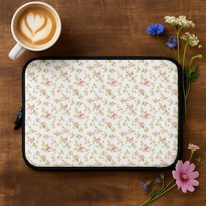 Può includere: Una custodia per laptop con un motivo floreale di piccole rose rosa e foglie verdi su sfondo bianco. La custodia ha un bordo nero e una cerniera nera. Una tazza di caffè con latte art e fiori sono accanto alla custodia.