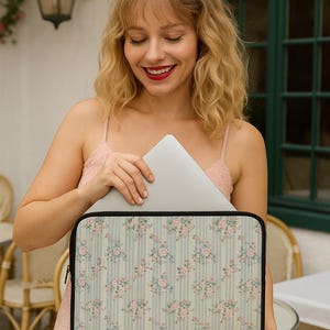 Può includere: Una custodia per laptop con un motivo floreale di rose rosa e strisce blu. La custodia ha un bordo nero e una chiusura a cerniera. Lo sfondo è un motivo a righe blu chiaro e bianco.
