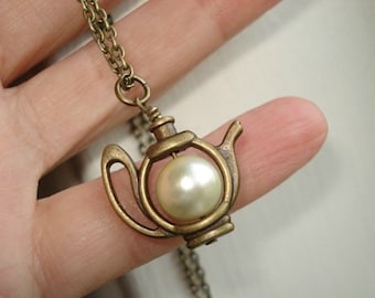 May include: A gold-tone teapot pendant necklace with a pearl in the center. The teapot has a delicate design and is a charming piece of jewelry.