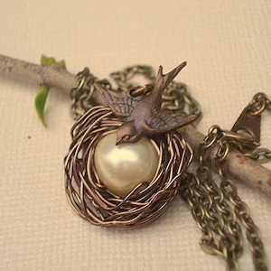 May include: A delicate necklace featuring a bird perched on a pearl inside a wire nest. The necklace is made of bronze-colored metal and has a simple chain.