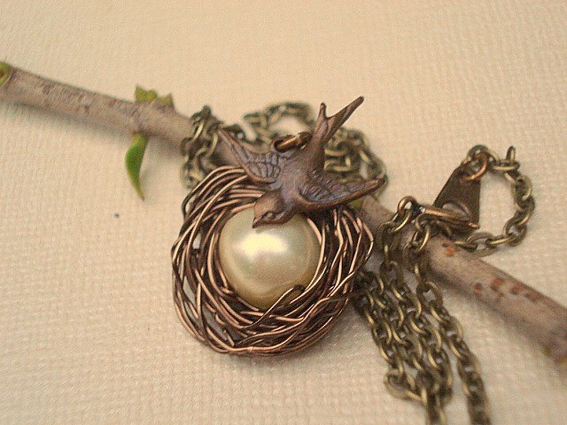 May include: A delicate necklace featuring a bird perched on a pearl inside a wire nest. The necklace is made of bronze-colored metal and has a simple chain.