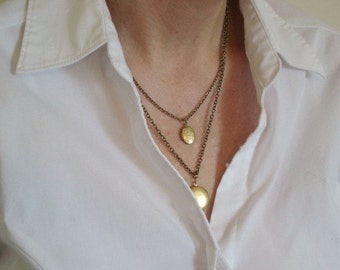 May include: A layered necklace set featuring two antique brass-toned chains. The shorter chain holds a small, oval-shaped locket with an embossed floral pattern, while the longer chain features a larger, smooth, round locket pendant.