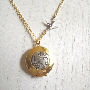 May include: Gold-tone necklace with a round locket pendant featuring a globe design. A small silver-tone bird charm is attached to the chain. The locket is hinged and appears to open. The necklace is displayed on a white wooden surface.