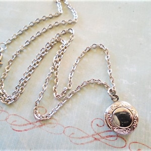 May include: A silver-colored locket necklace with a detailed, round pendant. The pendant and chain are made of a shiny metal. The necklace is displayed on a light blue surface with red decorative swirls.