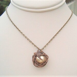 May include: A gold chain necklace with a pendant featuring a bird in a wire nest with a pearl in the center.