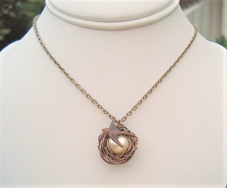 May include: A gold chain necklace with a pendant featuring a bird in a wire nest with a pearl in the center.