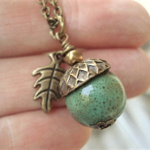 May include: A bronze-toned necklace featuring an acorn pendant with a green speckled ceramic body and a textured bronze cap. A bronze leaf charm hangs beside the acorn. The necklace is a nature-inspired accessory.