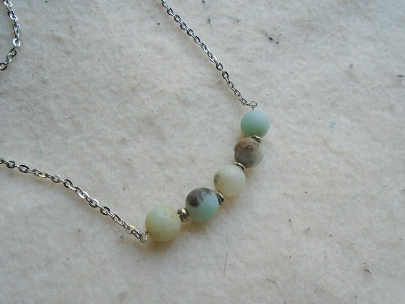 Puede incluir: Un collar de cadena plateada con una fila de cinco cuentas de piedras preciosas de color verde claro y blanco.