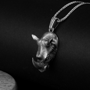 Può includere: Una collana con pendente color argento raffigurante una testa di rinoceronte dettagliata. Il pendente a forma di rinoceronte ha una superficie strutturata e un corno prominente. La catena della collana è a trama intrecciata e lo sfondo è nero.