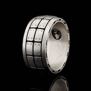 Peut inclure: Une bague en argent avec un design texturé composé d'une série de carrés avec de petits cercles autour des bords. La bague est marquée "925" sur le côté.