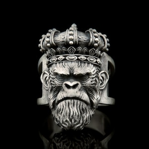 Peut inclure: Une bague en argent représentant une tête de singe sculptée en détail portant une couronne. Le singe a une expression sérieuse et une épaisse barbe.